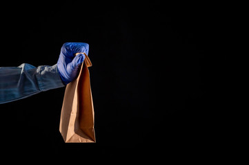 Female hand in a blue glove holds a brown paper bag on a black background. Safe food delivery to your home. A woman in a denim shirt holds out a cardboard bag to a customer.