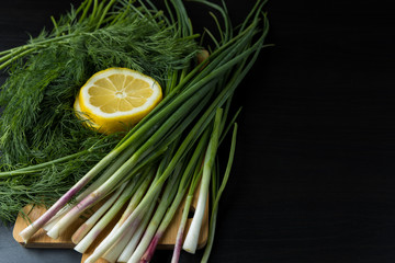 The smell of spring. Fresh spring greens for salads, green onions and dill, lemon slices on a wooden board and a dark wooden background. Top view, text area.