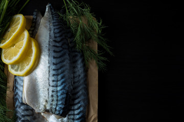 Fish products, commercial fish mackerel. Raw mackerel fillet with skin on a wooden board and on craft paper surrounded by green onions, dill and lemon slices. On a dark wooden background. Text area