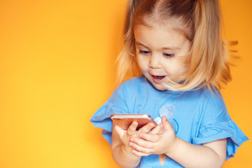  Cute little girl holding a smartphone and looking surprised with her mouth open, on an orange...