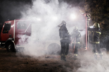 Obraz premium Team of professional firefighters with water hose in front of a firetruck with smoke in the air.