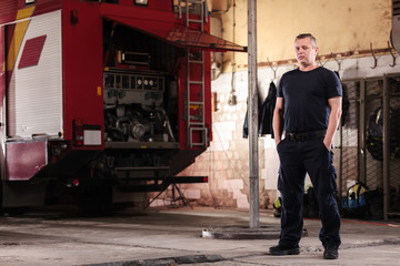 Fototapeta na wymiar Professional fireman portrait. Firefighter wearing uniform of shirt and trousers. Fire truck in the background.