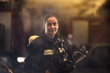 female firefighter portrait wearing full equipment and emergency rescue equipment. taking off oxygen mask after successful intervention. smoke and fire trucks in the background.