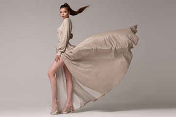 Woman in a long stylish evening dress posing in the studio. The train of the dress flies and...