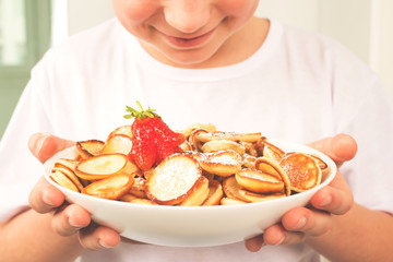 Happy child boy holding white bowl with healthy trendy mini pancakes cereal and strawberries. No...