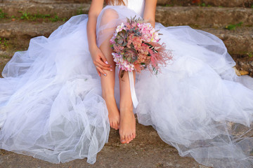 Naklejka na ściany i meble Young bride is sitting on the stone steps in a fluffy white skirt barefoot and holding a wedding delicate bouquet of succulents. Bride’s legs and hands are in a close-up view. Summer wedding day. 