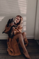 Girl in mustard yellow linen bathrobe sits on the floor. Blonde girl. Direct sunlight. Sunlight stripes. Holding cat. Pretty woman poses to the camera. Linen light bathrobe. Love and treat yourself.