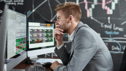 Securely invest to get profit. Portrait of successful young trader looking focused while sitting in front of multiple monitors in the office. Blackboard full of chart and data analyses in background.