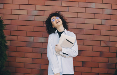 Caucasian woman with curly hair posing outside on a stone wall with eyeglasses holding a laptop