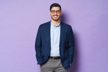 Young man wearing jacket, shirt and glasses, isolated on purple background, holding hands in pockets, looking straight at camera with happy smile