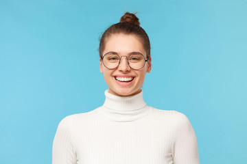 Headshot of happy teen girl wearing white turtleneck and eyeglasses, enjoying communication with mates and laughing at jokes, isolated on blue background