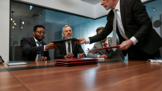 Discussing new poject. Mature businessman in formal wear giving folders to his multicultural colleagues while having a meeting in the office