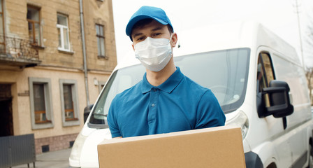 Fototapeta na wymiar Portrait of young courier in mask standing near delivery car and holding carton box