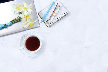 The desktop of a modern woman, home office. A computer, a cup of tea and spring flowers on a light table. Minimal concept of business, home comfort, top view, flat lay, place for text.