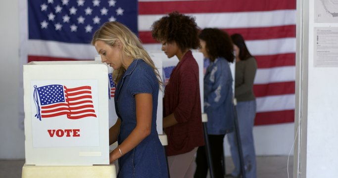 Four women of various demographics, young blonde woman in front, filling in ballots and casting votes in booths at polling station, US flag on wall at back