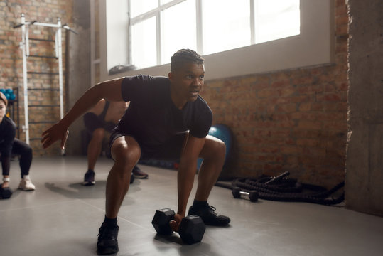 Train Hard, Live Better. Full Length Shot Of Athletic Man Lifting Dumbbell While Having Workout At Industrial Gym. Group Training, Teamwork Concept