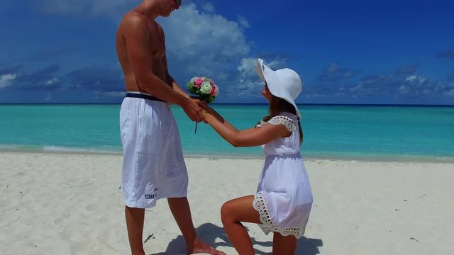 Woman kneeling on one knee while giving flowers to a man in the middle of the beach during daytime.