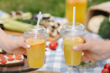 Photo of two hand hold orange lemonade in front of vegan picnic outdoors. Healthy food concept