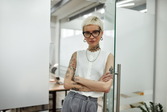 Successful business lady. Young beautiful blonde tattooed businesswoman with short haircut leaning on the glass door, keeping arms crossed and looking at camera while standing in modern office