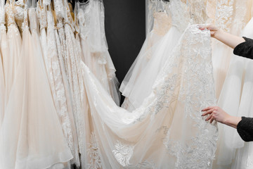 Bride touches luxurious white cream wedding dress on hangers. Dresses made of silk chiffon, tulle and lace. Luxury pearls and crystals pendants on the sleeves of a delicate color of a wedding dress