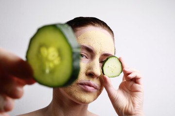 Headshot of Young woman with a face mask on holding a cucumber slice in front of one eye. Beauty...