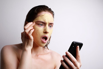 Close-up portrait of young caucasian woman with surprising face applying facial clay mask using her smartphone.
