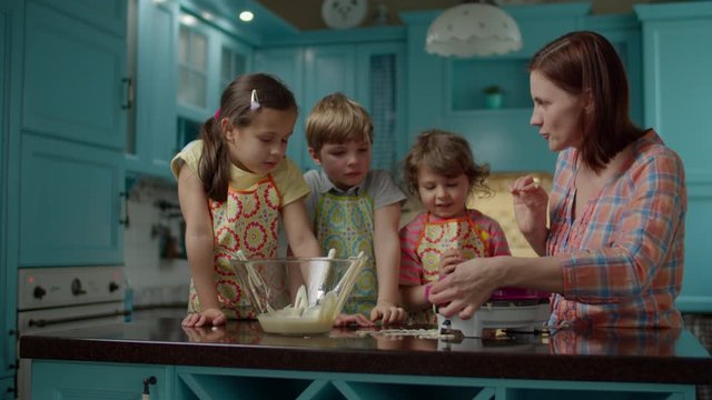Young 30s mother with three kids in aprons making waffles in waffle maker at home blue kitchen. Children putting dough into waffle maker with spoons. Cooking together. 