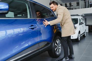 Visiting car dealership. Handsome bearded man is stroking his new car and smiling