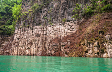 Wushan, Chongqing, China - May 7, 2010: Mini Three Gorges on Daning River. Closeup of multi-color browns and grays rocky cliff above emerald green water. Some green foliage.