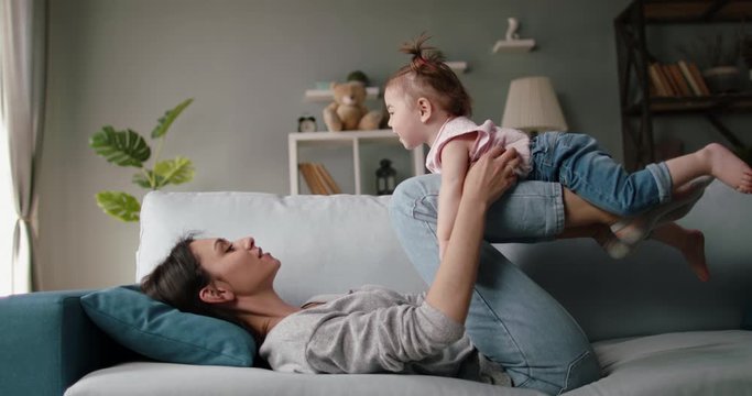 Active caucasian young mom and her little cute daughter playing at home, playing plane on couch. Joyful babysitter working at home - happy family, activity together concept 4k footage