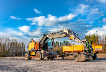Obraz premium Beautiful large yellow excavator with big wheels stands close to yellow service cart on new not yet ready road. Sunny evening with blue clouds, spring time, mixed forest at background. Side view