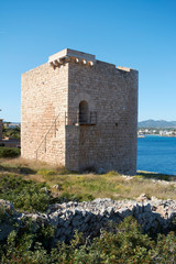 Mediterranean landscape on a sunny, foggy morning. Medieval tower in the foreground and small fishing village in the background. Porto Petro, Mallorca. Balearic Islands. Spain