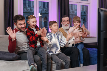 Attractive three generations of people as father, grandpa and grandchildren which sitting on the comfortable couch at home and enjoying leisure watching soccer match, shouting when team scored ball.