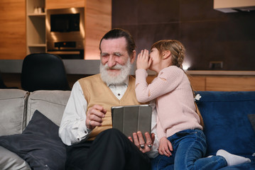 Slow motion of cute joyful 12-aged girl with funny pigtails which whispering on granddad's ear her secret while they sitting together on the sofa at home