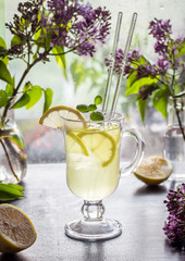 Refreshing cold yellow lemonade in a transparent cup and lilac in a vase are on the table