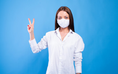 Portrait of an attractive young female doctor in white coat and mask, show two fingers