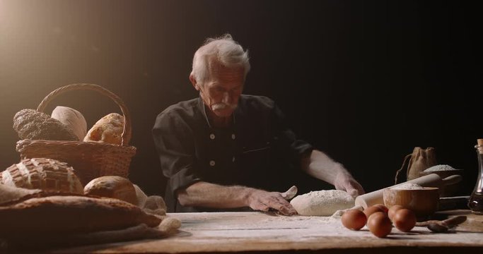 Professional mature caucasian baker putting flour on loaf of bread dough with passion, retired pensioneer enjoying his new hobby, isolated on black background 4k footage