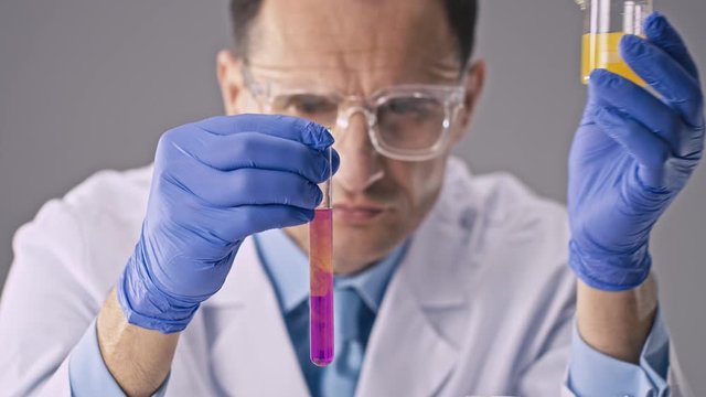 A chemist or laboratory assistant in a white coat and glasses in an innovative laboratory oversees the process of mixing multi-colored chemicals in test tubes. Scientific Innovation Research Concept