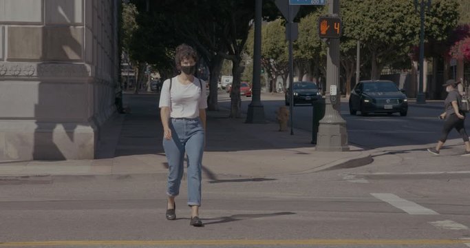 Woman Wearing a Face Mask Crosses Street Towards Camera in Downtown Los Angeles Intersection During Corona Virus Pandemic