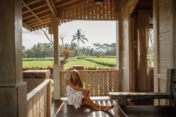 Attractive woman sitting on veranda of wooden house, relaxed summery atmosphere. Vacation on Bali, Ubud. Tropical view on background, palm tree and rice terraces