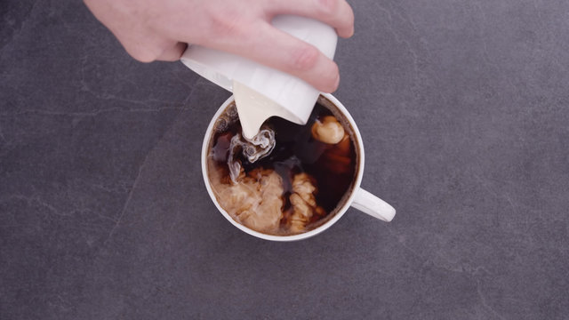 Top Overhead View Of Big Coffee Cup With A Plate Standing On Dark Stone. Cream Is Poured Into The Cup.