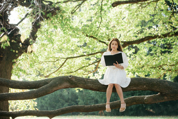 Beautiful asian woman in white dress sit on tree branch writing and thinking in the park with green leaves.