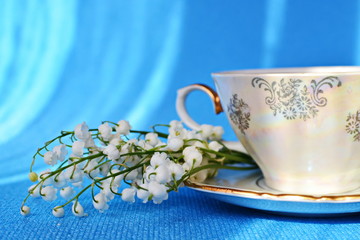 Part of porcelain pattern teacup with golden handle and bunch white flower of lily of the valley on saucer close up on blue background 