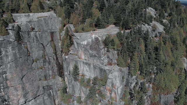 Man Standing On The Edge Of The Rocky Cliff In Mount Pinacle - Stunning Landscape On Top Of Mount Pinacle In Quebec, Canada With Lush Pine Trees- aerial drone shot