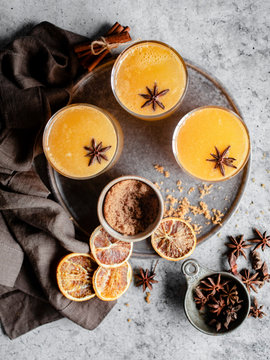 Overhead shot of three glasses of an orange spiced drink on a silver tray, garnished with star anise and surrounded by blood oranges, cinnamon sticks, a grey napkin, and a small bowl of brown sugar