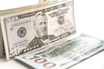 A United States paper currency fifty dollar bill stands on edge in front of a pile of other cash with the back side of a hundred dollar bill lays on another cash stack in front on a white background.