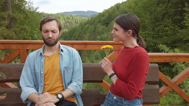 Cute woman sits next to man on a bench gives man to sniff dandelion, Man begins an allergic reaction and he begins to sneeze. Mountains on background