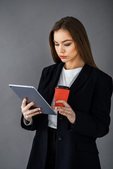 Young pretty girl in business style clothes stands on a gray background with a tablet and a cup of coffee in her hands. Business style