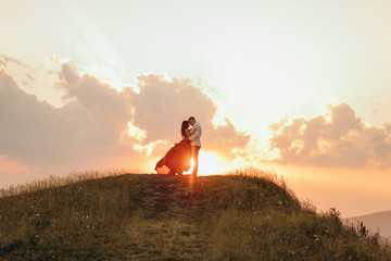 Happy couple stands in embrace and kisses each other on beautiful sunset in mountain. - obrazy, fototapety, plakaty