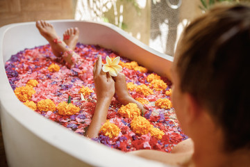 Concept of spa procedure. Dreamy young adult female taking bath with flowers petals, relaxing in bathroom, holding flower in her hands. Aromatherapy with flower petals